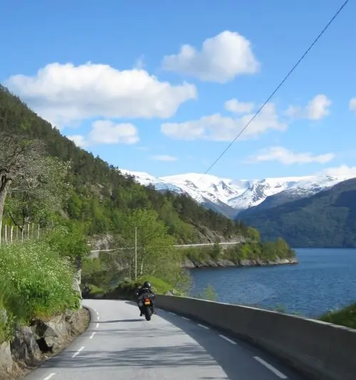 Motorrijder langs fjord met besneeuwde bergen tijdens motorreis Norwegian Dream, Noorwegen