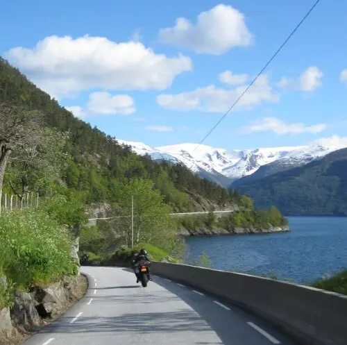 Motorrijder langs fjord met besneeuwde bergen tijdens motorreis Norwegian Shortcut, Noorwegen