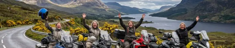 Vier enthousiaste motorrijdsters juichend op hun motoren langs een loch in de Schotse Highlands tijdens een motorvakantie Schotland