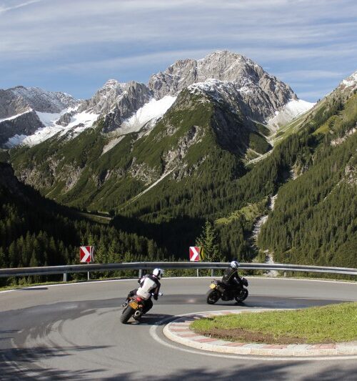 Motorrijders in een Alpenbocht met besneeuwde bergtoppen